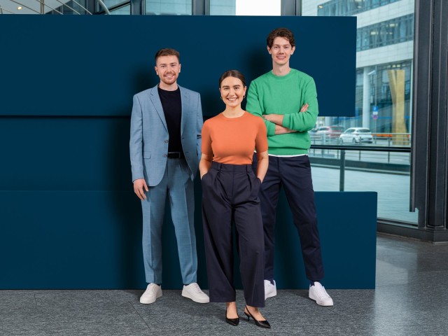 Drei Trainees stehen lächelnd vor einer dunkelblauen Wand.