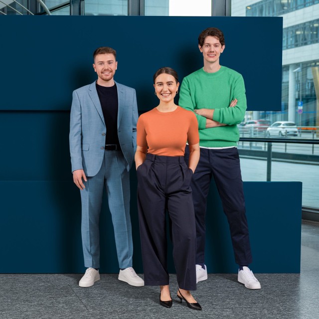 Drei Trainees stehen lächelnd vor einer dunkelblauen Wand.