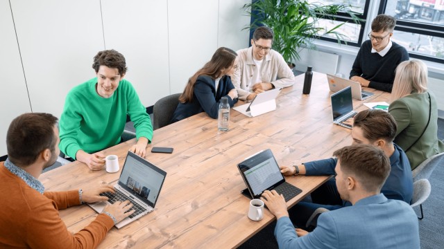 Personen arbeiten an einem großen Konferenztisch mit Laptops und Tablets.