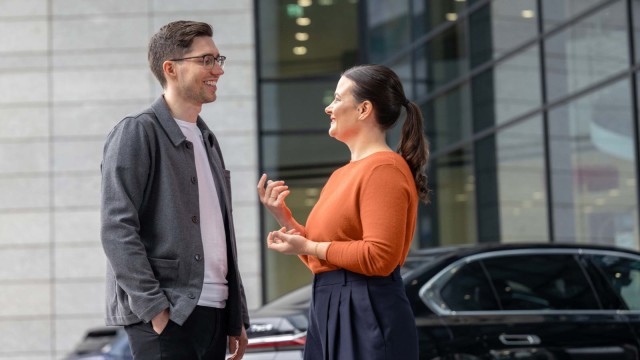 Zwei Personen unterhalten sich vor einem Gebäude neben einem schwarzen Auto.