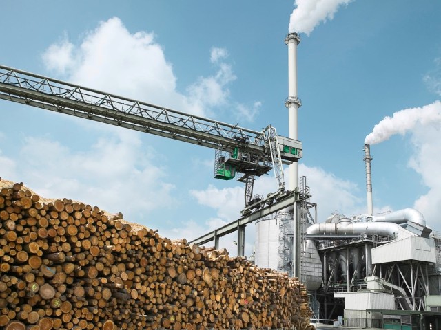 Ein industrielle Fabrik mit rauchenden Schornsteinen und einem Stapel Holzstämme im Vordergrund