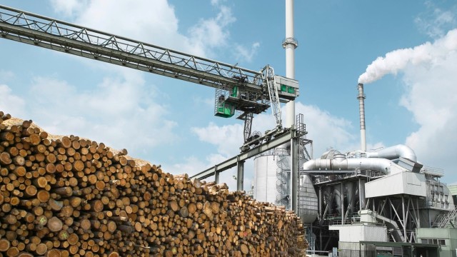 Ein industrielle Fabrik mit rauchenden Schornsteinen und einem Stapel Holzstämme im Vordergrund