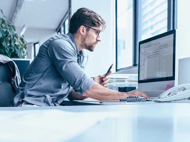 Mann mit Brille und hellblauem Hemd sitzt arbeitend am Schreibtisch und blickt auf einen Monitor